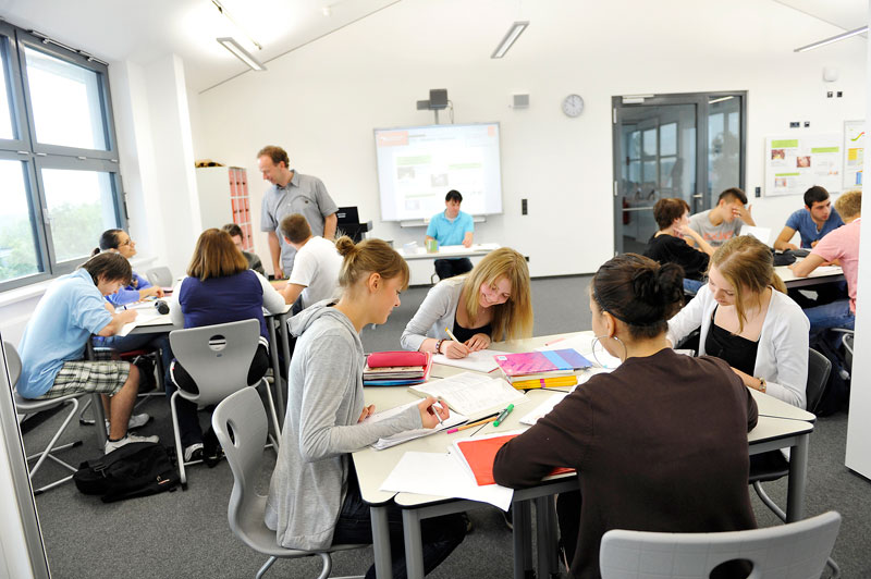 Mehrere Schülerinnen und Schüler sitzen in einem modernen Klassenzimmer an Tischen und arbeiten gemeinsam an Aufgaben. Ein Lehrer steht vorne am Raum neben einem interaktiven Whiteboard. Dies spiegelt eine Umgebung wider, in der sowohl Lehren als auch Lernen gefördert werden, was für Konzepte der Prävention und des Arbeitsschutzes relevant ist. Die helle Beleuchtung und die ergonomischen Möbel unterstützen eine positive Lernatmosphäre.