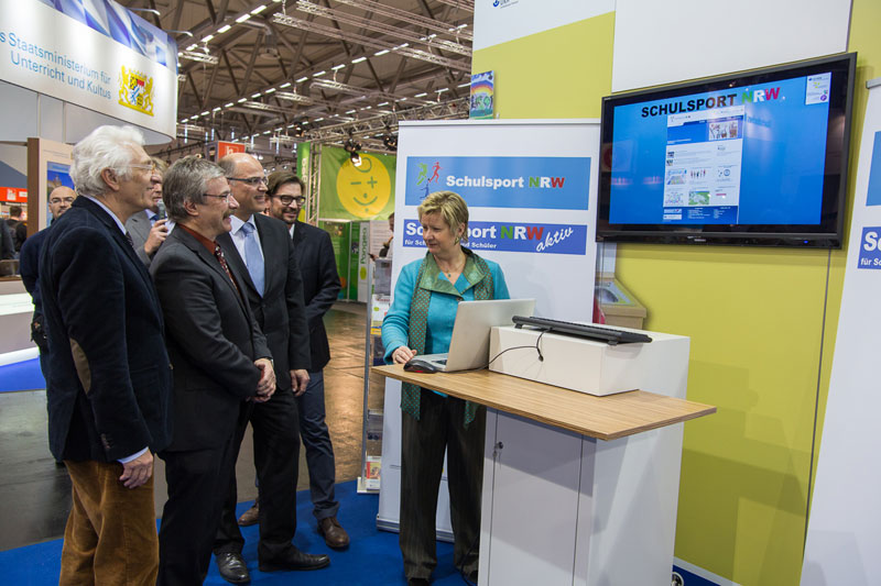 Eine Gruppe von sechs Personen steht vor einem Messestand von Schulsport NRW. Eine Frau zeigt auf einem Laptop eine Präsentation, die auch auf einem großen Bildschirm zu sehen ist. Die Gruppe, bestehend aus Männern in Anzügen und einem Mann in einem blauen Samtjackett, beobachtet interessiert die Präsentation. Im Hintergrund sind weitere Messestände und Dekorationen zu erkennen. Der Kontext deutet auf Arbeitsschutz und Prävention im Schulsport hin.