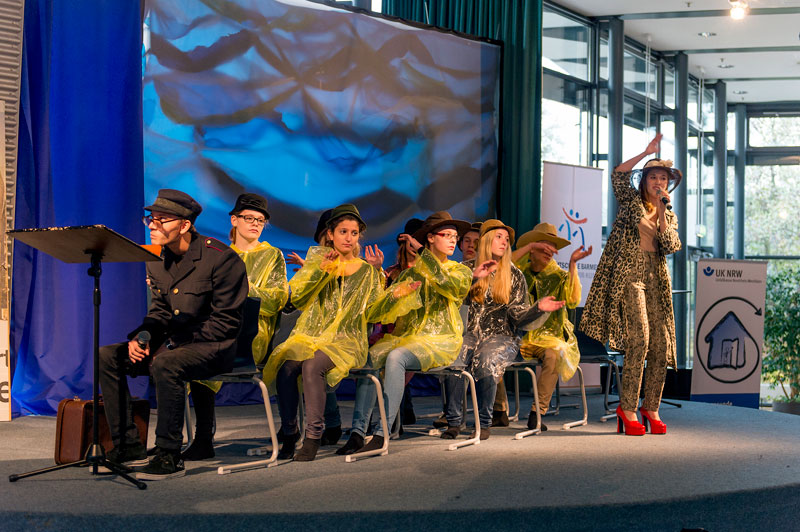 Eine Theatergruppe führt auf einer Bühne ein Stück auf, das sich mit Arbeitsschutz thematisch befasst. Sechs Personen sitzen auf Stühlen und tragen gelbe Regenmäntel und schwarze Hüte, was möglicherweise die Darstellung von Sicherheitsaspekten in der Arbeit symbolisiert. Eine Person steht rechts und spricht mit einem Mikrofon, gestikuliert und trägt ebenfalls einen Hut. Ein Mann in dunkler Kleidung liest von einem Podium ab. Im Hintergrund sind Banner der Unfallkasse NRW sichtbar, was auf ein Event im Kontext von Prävention und Arbeitsschutz hinweist. Die Szenerie befindet sich in einem gut beleuchteten Raum mit großen Fenstern.