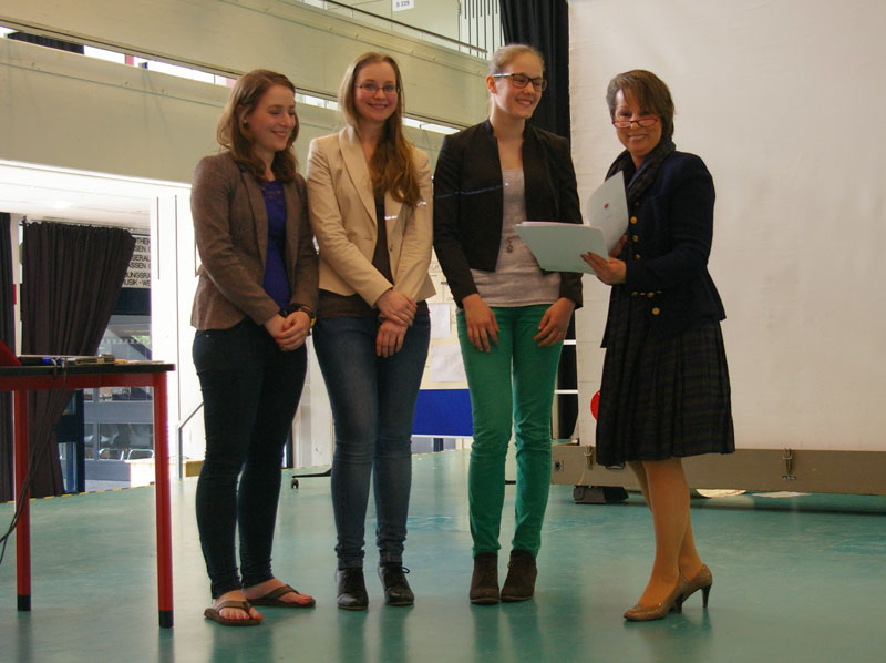 Drei junge Frauen stehen nebeneinander, lächelnd auf einer Bühne. Eine vierte Person überreicht ihnen eine Urkunde. Die Szene wirkt wie eine Preisverleihung. Im Hintergrund sind ein Tisch, ein Vorhang und eine große Leinwand zu sehen. Der Kontext scheint mit Arbeitsschutz und Prävention zu tun zu haben, möglicherweise eine Veranstaltung der Unfallkasse NRW.