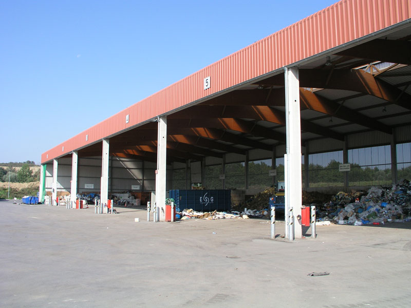 Eine große, offene Lagerhalle mit fünf deutlich sichtbaren Eingängen. Unter der Überdachung sind Recyclingabfälle und Müll verteilt. Blaue Container und verschiedene Abfall- und Recyclingmaterialien sind sichtbar. Das Bild zeigt den Kontext von Arbeitsumgebung und Sicherheitsaspekten im Bereich Abfallmanagement und Recycling. Der Himmel ist klar und das Gelände sauber, was auf eine geregelte, überwachte Umgebung hinweist.