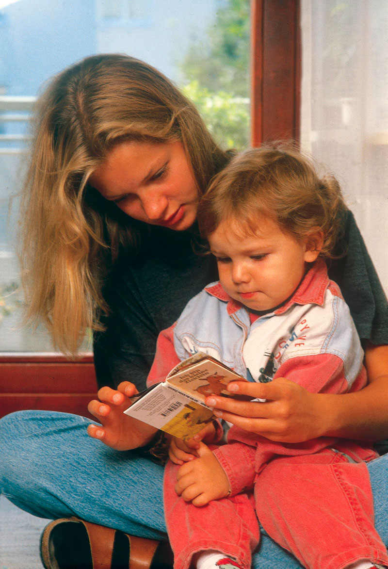 Eine blonde Frau sitzt mit einem Kleinkind auf dem Schoß und liest ihm ein Bilderbuch vor. Beide sind in legere Kleidung gekleidet und sitzen in einem hellen Raum nahe eines großen Fensters. Die Szene deutet auf Bildungsförderung und elterliche Fürsorge hin, was im Kontext der Unfallkasse NRW auf die Wichtigkeit der frühkindlichen Bildung und Prävention hinweist.