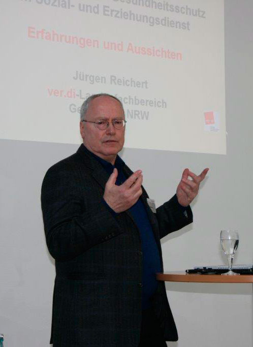 Ein Mann mittleren Alters hält einen Vortrag, gestikulierend vor einer Projektion. Auf der Leinwand ist eine Präsentation zu Gesundheits- und Arbeitsschutz im Sozial- und Erziehungsdienst mit dem Titel 'Erfahrungen und Aussichten' zu sehen. Der Referent trägt einen dunkelgrauen Anzug und ein blaues Hemd. Vor ihm auf einem Tisch steht ein Glas mit Wasser und ein Laptop.