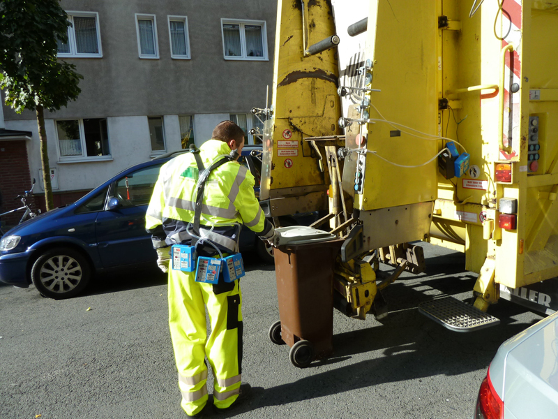 Ein Müllabfuhrmitarbeiter in leuchtend gelber Schutzkleidung leert einen braunen Mülleimer in einen gelben Müllabfuhr-LKW auf einer Straße. Der Mitarbeiter trägt eine Sicherheitsweste und Arbeitshandschuhe, was auf Maßnahmen zur Unfallverhütung hinweist. Im Hintergrund sind ein Auto und ein Gebäude zu sehen, die typische städtische Umgebung hinzufügen.
