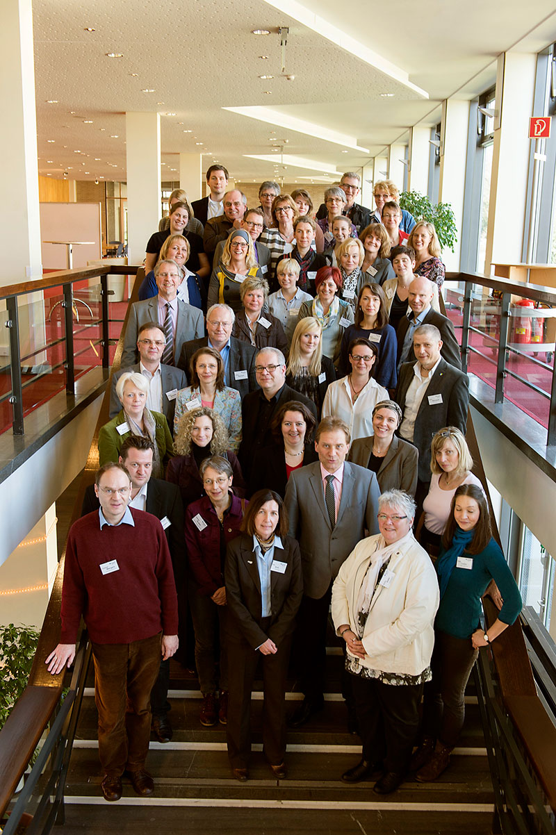 Eine große Gruppe von Menschen posiert auf einer breiten Treppe in einem modernen Konferenzgebäude. Die Personen sind in mehreren Reihen angeordnet und blicken direkt in die Kamera. Einige tragen Namensschilder, die Umgebung ist hell erleuchtet mit großen Fenstern und Pflanzen im Hintergrund. Dieses Bild könnte in einem Kontext von Arbeitsschutz und Prävention verwendet werden, um eine Veranstaltung oder ein Treffen der Unfallkasse NRW darzustellen.