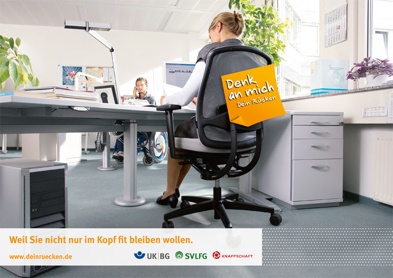 Eine Frau sitzt an einem Schreibtisch und arbeitet am Computer. Eine weitere Person im Rollstuhl ist im Hintergrund zu sehen. Auf der Rückseite des Bürostuhls der Frau befindet sich ein Schild mit der Aufschrift 'Denk an mich. Dein Rücken'. Der Raum ist gut beleuchtet mit großen Fenstern im Hintergrund. Im Vordergrund ist ein Banner mit den Logos mehrerer Organisationen, darunter UK IBG, SVLFG und Knappschaft, und dem Text 'Weil Sie nicht nur im Kopf fit bleiben wollen.'
