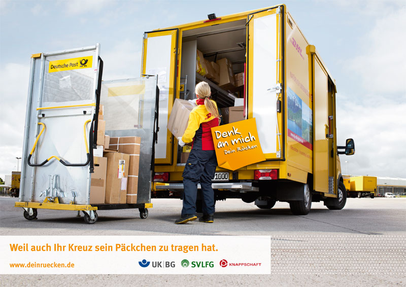 Eine Mitarbeiterin der Deutschen Post belädt einen gelben Lieferwagen mit Paketen. Der Lieferwagen steht auf einem Lieferhof mit mehreren Gebäuden im Hintergrund. Auf dem Lieferwagen steht 'Denk an mich - Dein Rücken'. Die Szene betont die Wichtigkeit von sicherem Heben und Transportieren von schweren Gegenständen zur Vermeidung von Rückenproblemen. Die Botschaft 'Weil auch Ihr Kreuz sein Päckchen zu tragen hat' hebt das Thema Rückengesundheit hervor. Unten im Bild sind Logos von Unfallkassen zur Arbeitssicherheit und Prävention.
