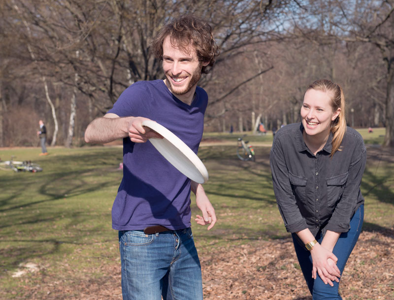Ein Mann in einem lila T-Shirt wirft eine Frisbee im Park, während eine Frau in einem grauen Hemd daneben steht und lächelt. Im Hintergrund sind Bäume ohne Laub und eine Person, die selbstständig Fahrrad fährt. Die Szene vermittelt Freude und Bewegung an der frischen Luft, was zur Prävention und Förderung der Gesundheit beiträgt.