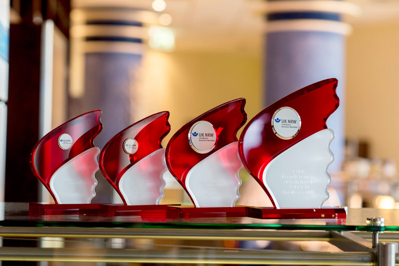 Das Bild zeigt vier rote Glastrophäen mit geschwungenen Formen, die auf einem Tisch nebeneinander stehen. Jede Trophäe trägt das Logo der Unfallkasse NRW und eine Gravur, die den jeweiligen Anlass oder die Leistung würdigt. Im Hintergrund ist ein unscharfer Bereich zu erkennen, der eine innenarchitektonische Szenerie andeutet. Die Trophäen symbolisieren Anerkennung und Erfolg im Bereich Arbeitsschutz und Prävention.