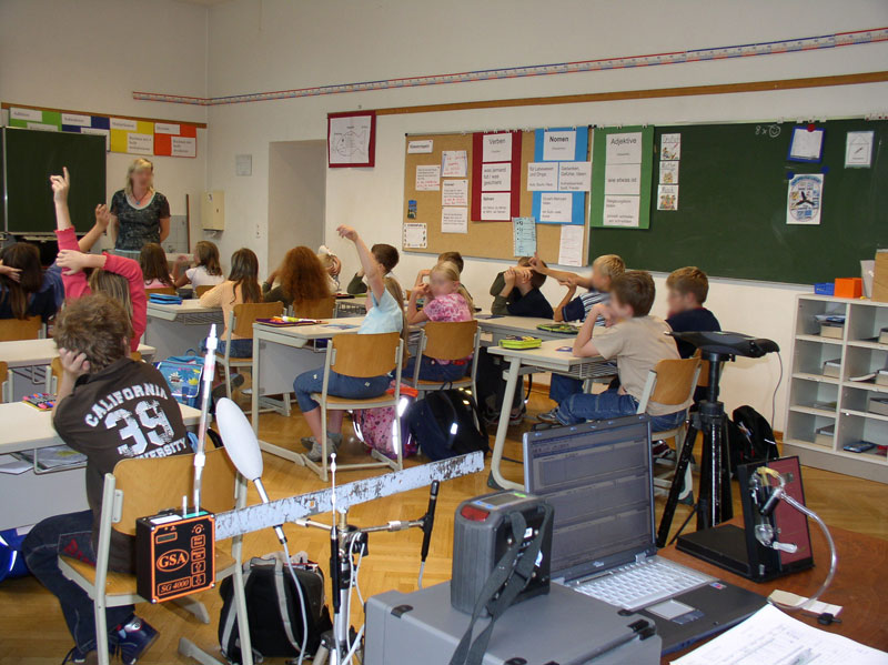 Ein Klassenzimmer mit mehreren Schülern, die an Tischen sitzen und die Hände heben. Eine Lehrkraft steht vor der Tafel. An der Wand hängen Lernplakate und an der Tafel sind Notizen erkennbar. Der Raum ist hell beleuchtet. Im Vordergrund stehen technische Geräte, die möglicherweise zur Unterstützung des Unterrichts oder für Sicherheitszwecke dienen. Das Bild könnte im Kontext der Unfallkasse NRW auf Arbeitsschutzmaßnahmen in Bildungseinrichtungen hinweisen.