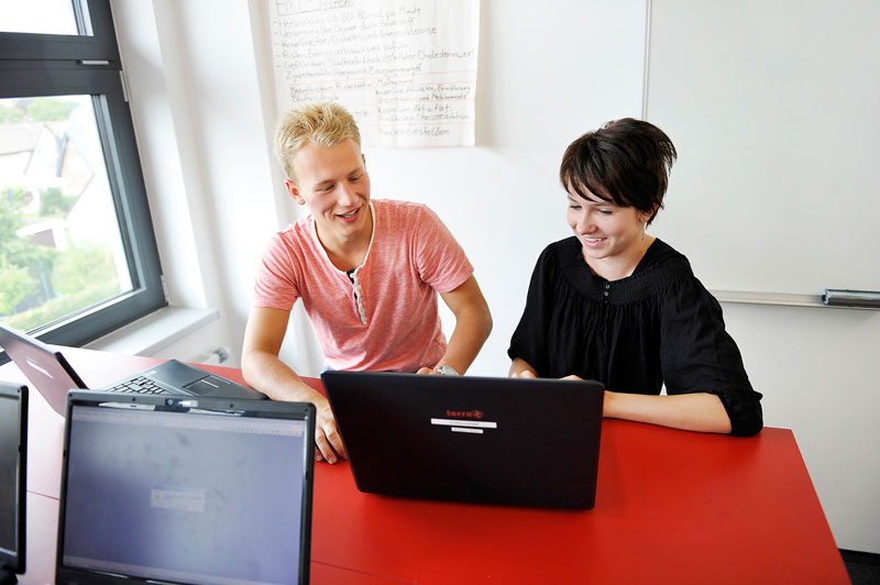 Ein junger Mann und eine junge Frau sitzen an einem roten Tisch und arbeiten gemeinsam an einem schwarzen Laptop. Im Hintergrund sind ein Fenster und eine beschriebene Tafel oder ein Poster zu sehen. Der Raum wirkt wie ein moderner Schulungsraum, der auf Teamarbeit und Zusammenarbeit ausgerichtet ist. Diese Umgebung fördert den Austausch und die Sicherheit am Arbeitsplatz, im Kontext der Unfallkasse NRW für Arbeitsschutz und Prävention.