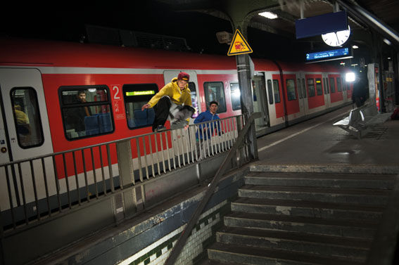 Das Bild zeigt zwei junge Personen, die an einem Bahnsteig über ein Geländer springen. Eine Regionalbahn steht im Hintergrund. Eine Person trägt einen gelben Hoodie und eine rote Mütze. Das Schild in der Nähe warnt vor Gefahren. Das Bild veranschaulicht unachtsames Verhalten im öffentlichen Raum, das sicherheitsrelevant ist im Kontext von Arbeitsschutz und Prävention.