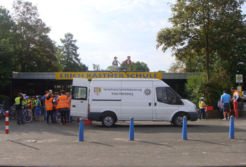Ein weißer Lieferwagen der Verkehrssicherheitsberatung Kreis Heinsberg steht vor der Erich Kästner Schule. Mehrere Erwachsene in orangefarbenen Warnwesten und Kinder befinden sich neben dem Fahrzeug. Auf dem Vordach der Schule stehen zwei Figuren, die Radfahrer darstellen. Einige Bäume und ein Haltestellenschild sind im Hintergrund zu sehen. Der Kontext deutet auf eine Veranstaltung zur Verkehrssicherheit und Prävention hin.