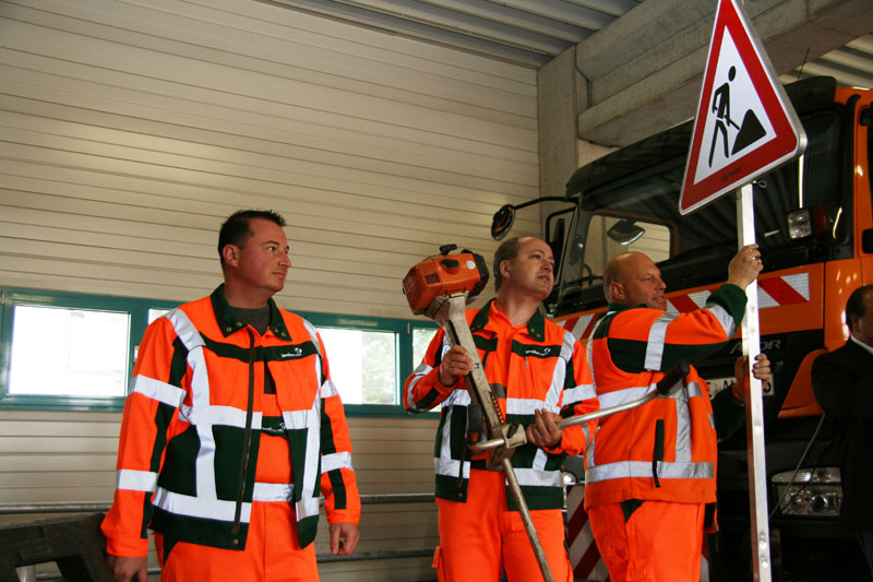 Drei Männer in orangefarbener Warnkleidung stehen in einer Werkshalle. Zwei halten Sicherheitsutensilien: Einer einen Vorschlaghammer, der andere ein Baustellenschild. Im Hintergrund ist ein großes Fahrzeug sichtbar. Diese Szene betont das Thema Arbeitsschutz und Prävention, relevant für die Unfallkasse NRW. Die Umgebung weist typische Sicherheitsausrüstungen auf, die für Straßenbauarbeiten genutzt werden.