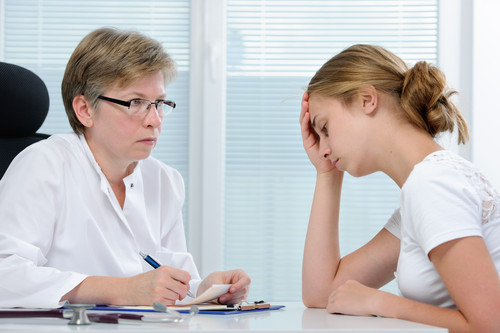 In einem hellen Sprechzimmer sitzt links eine Ärztin in weißem Kittel an einem Schreibtisch. Sie hält einen Schreibblock und einen Stift. Rechts sitzt eine Patientin im weißen T-Shirt und stützt ihren Kopf nachdenklich mit der Hand ab. Die Ärztin schaut aufmerksam zur Patientin. Das Bild thematisiert Arbeitsschutz und Prävention, indem es professionelle, einfühlsame Kommunikation im Gesundheitsumfeld symbolisiert, was für die Unfallkasse NRW relevant ist.