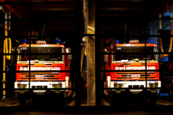 Zwei rot-weiße Feuerwehrautos sind in einer Feuerwehrstation hinter Metallgittern geparkt. Die Front der Fahrzeuge zeigt nach vorne, und auf beiden ist das Wort 'FEUERWEHR' zu lesen. Die Umgebung wirkt ordentlich und bereit für einen möglichen Einsatz, im Einklang mit den Arbeitsschutz- und Präventionsrichtlinien der Unfallkasse NRW.