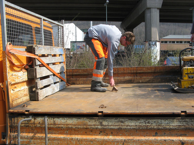 Ein Mann in Arbeitskleidung mit orangefarbenen Sicherheitsstreifen steht auf der Ladefläche eines LKWs unter einer Brücke. Er bückt sich, um den Laderaum sauber zu machen. Auf der Ladefläche befinden sich ein gesichertes Palettenstück und eine Baumaschine. Die Szene thematisiert Arbeitsschutz und Prävention bei LKW-Arbeiten im Kontext der Unfallverhütung.