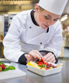Eine Köchin in weißer Uniform und Kochmütze steht in einer professionellen Küche und dekoriert einen Teller mit frischem Gemüse. Der Fokus liegt auf der genauen und hygienischen Zubereitung von Speisen, was den Wert von Arbeitsschutz und Prävention in gastronomischen Berufen unterstreicht. Die Küche im Hintergrund ist sauber und gut ausgestattet, was auf professionelle Standards hinweist.
