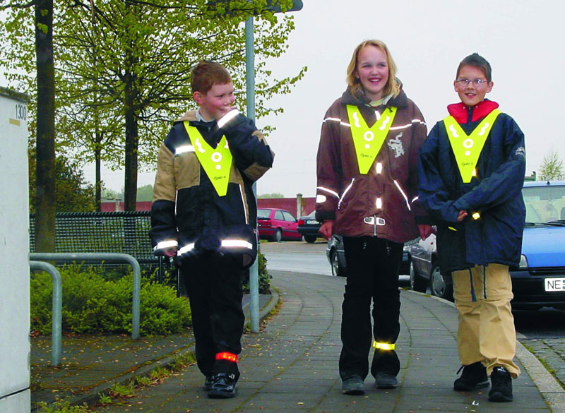 Drei Kinder gehen auf einem Bürgersteig. Sie tragen reflektierende Warnwesten über ihrer Kleidung, die zur besseren Sichtbarkeit im Straßenverkehr beitragen. Der Bürgersteig befindet sich neben einer Straße, auf der Autos geparkt sind. Im Hintergrund sind Bäume zu sehen. Dieses Bild verdeutlicht die Bedeutung von präventiven Sicherheitsmaßnahmen, insbesondere für Kinder im Straßenverkehr.
