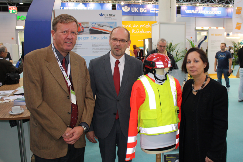 Drei Personen stehen nebeneinander neben einer Schaufensterpuppe mit auffälliger Schutzkleidung, bestehend aus neongelber Jacke und rotem Helm. Der Hintergrund zeigt einen Messestand der Unfallkasse NRW, der sich auf Arbeitsschutz und Prävention konzentriert. Im Hintergrund sind Informationsbanner und Pflanzen sichtbar, die eine professionelle und informative Atmosphäre schaffen.