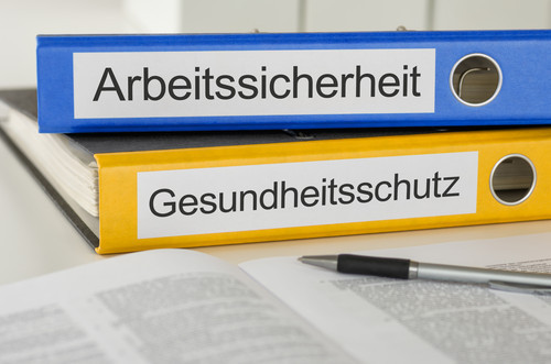 Zwei Ordner in blau und gelb mit den Beschriftungen 'Arbeitssicherheit' und 'Gesundheitsschutz' liegen übereinander auf einem Tisch. Im Vordergrund befindet sich ein aufgeschlagenes Buch, daneben liegt ein Kugelschreiber. Das Bild symbolisiert organisatorische Aspekte des Arbeitsschutzes und Gesundheitsschutzes, die für Prävention und Sicherheit am Arbeitsplatz wichtig sind.