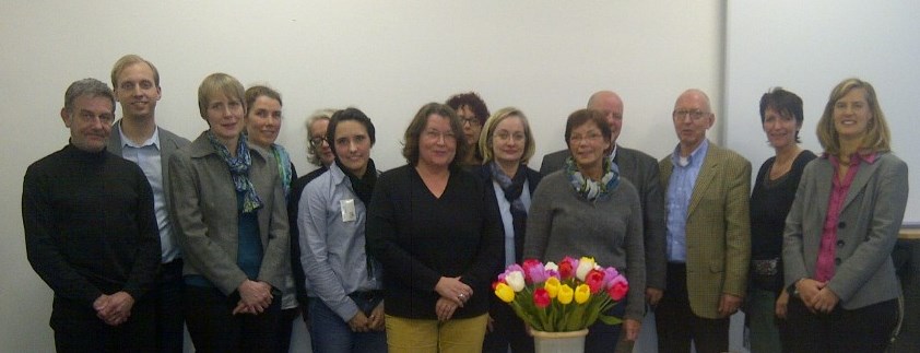 Eine Gruppe von elf Personen steht in zwei Reihen hinter einem Tisch mit einem bunten Blumenstrauß. Der Kontext deutet auf ein Meeting oder eine Veranstaltung hin, möglicherweise im Rahmen der Unfallkasse NRW, die sich mit Arbeitsschutz und Prävention befasst. Alle Personen lächeln oder blicken freundlich, einige tragen Blazer und sind unterschiedlich gekleidet, was auf eine förmliche, aber nicht allzu strenge Kleiderordnung hinweist.