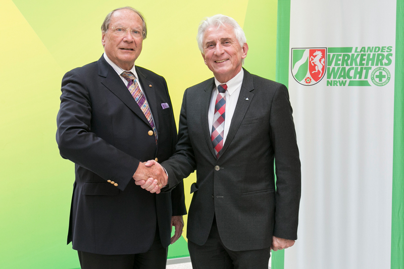 Zwei ältere Männer in Anzügen stehen nebeneinander und schütteln die Hände. Sie stehen vor einem Hintergrund mit dem Logo der Landesverkehrswacht NRW, einer Organisation, die sich auf Verkehrssicherheit und Prävention konzentriert. Der Kontext deutet auf Zusammenarbeit oder ein formelles Treffen im Bereich der Verkehrssicherheitsprävention hin.