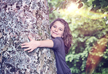 Ein Kind mit braunen Haaren umarmt in einem Wald einen großen Baumstamm voller Flechten. Sonnenstrahlen dringen durch das Blätterdach, während das Kind ein glückliches Lächeln zeigt. Das Bild vermittelt eine Botschaft von Naturverbundenheit und Umweltschutz, was im Kontext der Unfallkasse NRW auf die Bedeutung von Prävention und Arbeitsschutz in natürlichen Umgebungen hinweist.