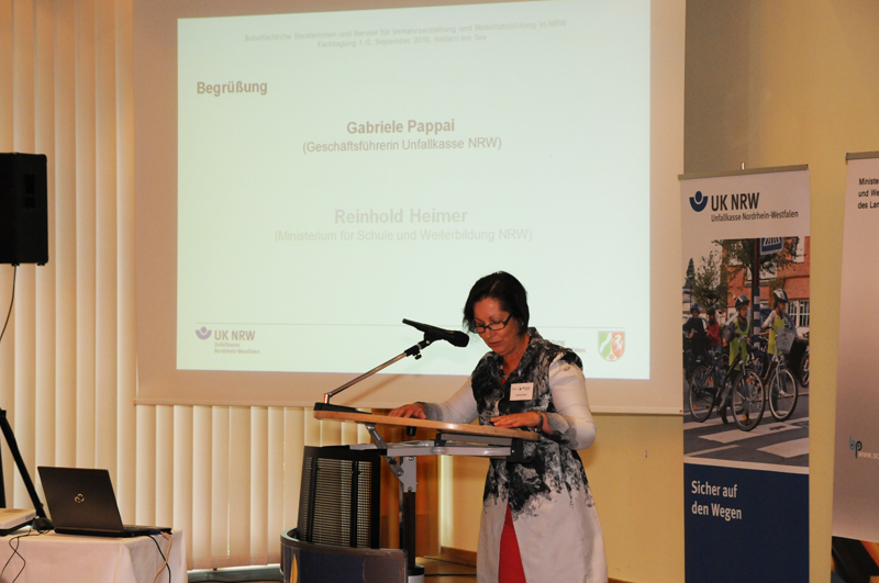 Eine Frau hält einen Vortrag bei einer Veranstaltung der Unfallkasse NRW. Im Hintergrund ist eine Projektionsfläche mit einer Präsentation, die die Begrüßung durch Gabriele Pappai von der Unfallkasse NRW und Reinhold Heimer vom Ministerium für Schule und Weiterbildung NRW zeigt. Rechts im Bild befinden sich ein Roll-up mit dem Logo der Unfallkasse NRW und ein Bild von Menschen, die auf einem Weg unterwegs sind. Der Raum ist schlicht gehalten mit Vorhängen und einem Pult mit Mikrofon.