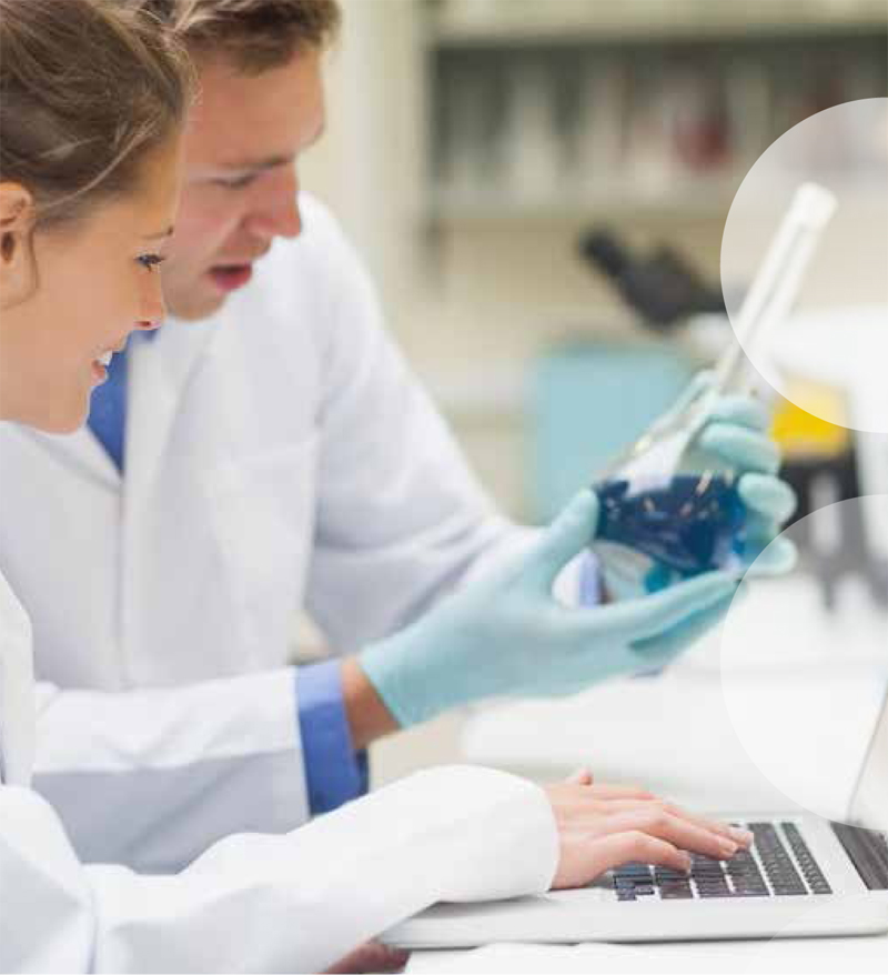 Zwei Laboranten in weißen Kitteln führen eine Analyse durch. Eine Frau tippt auf einem Laptop, während ein Mann ein Becherglas mit blauer Flüssigkeit hält. Im Hintergrund ist Laborgeräte unscharf zu sehen. Der Fokus liegt auf der Zusammenarbeit und Sicherheitskleidung, die für Arbeitsschutz wichtig ist.