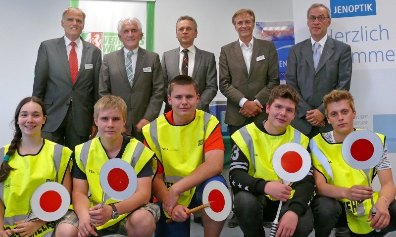Eine Gruppe von elf Personen bei einer Veranstaltung der Unfallkasse NRW. Im Hintergrund stehen fünf Männer in Anzügen, die vor Roll-Up-Bannern posieren. Im Vordergrund sitzen sechs junge Erwachsene in gelben Warnwesten, die rote und weiße Schilder halten. Die Veranstaltung steht im Kontext von Arbeitsschutz und Prävention.