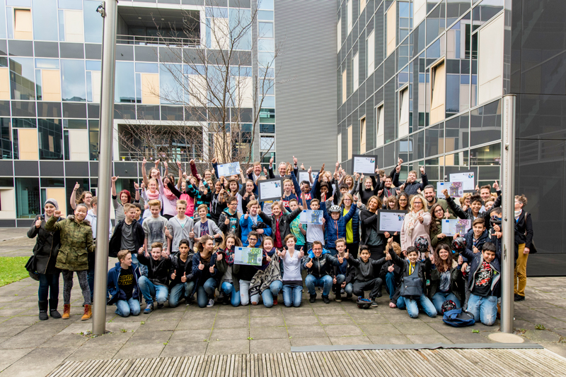 Eine große Gruppe unterschiedlicher Menschen, darunter Jugendliche und Erwachsene, versammelt sich vor einem modernen Bürogebäude. Sie halten stolz Zertifikate in den Händen und lachen in die Kamera. Die Umgebung ist städtisch mit einem gepflasterten Platz und Grünflächen. Die Veranstaltung könnte im Zusammenhang mit einem Sicherheits- oder Präventionsprojekt der Unfallkasse NRW stehen, was durch die fröhliche Atmosphäre und die Präsentation von Auszeichnungen unterstrichen wird.