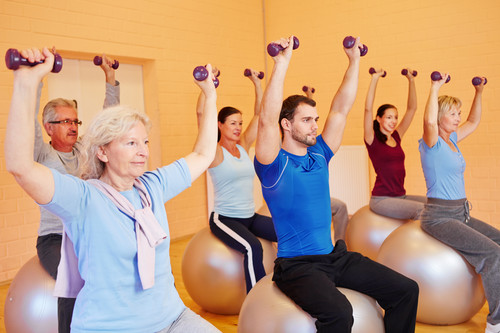 Sechs Personen verschiedenen Alters sitzen auf Gymnastikbällen in einem Fitnessraum. Sie halten leichte Hanteln in die Luft, während sie eine Übung durchführen. Der Fokus liegt auf Bewegung und Prävention am Arbeitsplatz, möglicherweise als Teil eines Programms der Unfallkasse NRW zur Förderung von Gesundheit und Sicherheit.