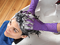 Eine Person mit lila Handschuhen wäscht die Haare der Kundin an einem Friseurwaschbecken. Die Kundin sitzt in einem Stuhl mit einem blauen Umhang, während der Schaum im Haar verteilt wird. Der Holzboden und der Wasserhahn sind sichtbar. Der Kontext bezieht sich auf Arbeitsschutz und Hygiene im Friseurgewerbe, wie von der Unfallkasse NRW thematisiert.