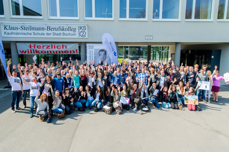 Eine große Gruppe von Menschen, größtenteils junge Erwachsene, steht vor dem Klaus-Steilmann-Berufskolleg in Bochum. Sie halten Plakate und Banner, auf einem ist 'Herzlich willkommen!' zu lesen. Viele heben die Arme und lächeln in die Kamera. Das Gebäude im Hintergrund trägt den Namen der Schule in großen Buchstaben. Die Veranstaltung scheint eine Willkommensveranstaltung oder ein Event zur Förderung von Gemeinschaft und Arbeitsschutz zu sein, was für die Unfallkasse NRW im Bereich Prävention relevant ist.