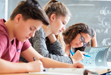 Drei Schüler sitzen in einem Klassenzimmer an einem Tisch und sind in ihre Aufgaben vertieft. Zwei Mädchen und ein Junge schreiben in ihre Hefte, während sie an einem Holzschreibtisch sitzen. Im Hintergrund ist eine Schultafel zu sehen. Dieses Bild vermittelt einen Fokus auf konzentriertes Lernen und Bildung, was relevante Aspekte der Arbeitsschutz- und Präventionsstrategien der Unfallkasse NRW, wie sichere und fördernde Lernumgebungen, unterstützen kann.