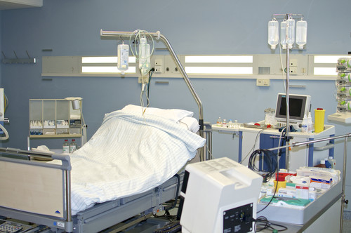 Ein Krankenhauszimmer mit einem Pflegebett, umgeben von medizinischem Equipment. Am Bett hängt ein Infusionsständer mit mehreren Beuteln. Rechts von dem Bett stehen ein Überwachungsmonitor und diverse medizinische Geräte sowie Verbrauchsmaterialien auf einem Tisch. Der Raum ist in hellen Farben gestaltet und dient der Überwachung und Erholung von Patienten, was im Kontext der Unfallkasse NRW auf Arbeitssicherheit und Schutzmaßnahmen hinweist.