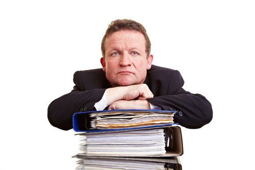 Ein Mann in Anzug lehnt mit verschränkten Armen auf einem hohen Stapel Aktenordner. Der Hintergrund ist weiß. Der Ausdruck des Mannes ist nachdenklich. Die Szene vermittelt Themen wie Büroarbeit, Dokumentation und Verwaltung, relevante Aspekte für den Arbeitsschutz und die Prävention im Kontext der Unfallkasse NRW.