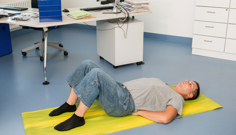 Ein Mann liegt auf einer gelben Gymnastikmatte auf dem Boden eines Büros. Er trägt ein graues T-Shirt, blaue Jeans und schwarze Socken. Seine Hände sind unter seinem unteren Rücken platziert, während er auf dem Rücken liegt. Im Hintergrund steht ein Bürotisch mit einem Stuhl, Computer, Akten und Papieren. Die Szene vermittelt die Bedeutung von Bewegung und Ergonomie am Arbeitsplatz zur Unfallprävention und Gesundheitsförderung.