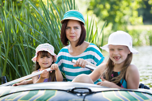 Eine Frau und zwei Kinder sitzen in einem Schlauchboot, das auf einem ruhigen Fluss fährt. Die Frau trägt ein gestreiftes blau-weißes T-Shirt und einen türkisfarbenen Hut. Beide Kinder haben Sommerhüte auf und lächeln. Im Hintergrund sind hohe Gräser und Bäume zu sehen. Diese Szene kann auf Präventionsmaßnahmen wie Tragen von Schwimmwesten für Sicherheit auf dem Wasser hinweisen.