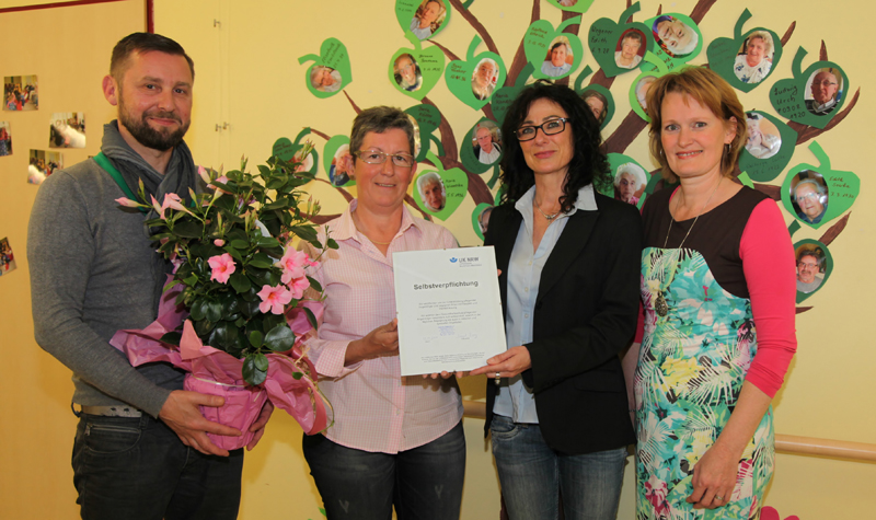 Vier Personen bei der Übergabe einer Urkunde und eines Blumenstraußes.