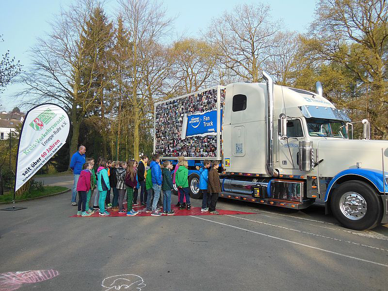 Eine Gruppe von Kindern steht in einer Reihe vor einem großen weißen Truck, der das Logo der Unfallkasse NRW trägt. Der Truck befindet sich auf einem Parkplatz, umgeben von Bäumen. Ein Erwachsener spricht mit den Kindern, die in bunter Freizeitkleidung gekleidet sind. Der Truck hat ein Banner mit einem Präventionsslogan. Links im Bild steht ein Werbebanner der Unfallkasse NRW. Der sonnige Tag unterstützt die freundliche Atmosphäre der Veranstaltung, die dem Zweck der Verkehrssicherheit und Prävention dient.