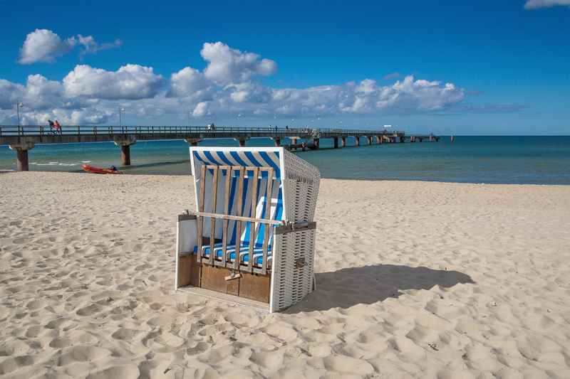 Im Vordergrund steht ein einzelner Strandkorb mit blauen und weißen Streifen auf feinem Sand. Im Hintergrund erstreckt sich ein Pier ins Meer unter einem leicht bewölkten Himmel. Die Szene wirkt ruhig und einladend, ideal für Erholung und Entspannung am Strand. Diese Umgebung kann als Erholungsmaßnahme im Sinne der Unfallkasse NRW betrachtet werden, die Arbeitsschutz und Prävention durch Regeneration und Pausen fördert. Menschen gehen auf dem Pier spazieren, der zum Meer hinausführt.
