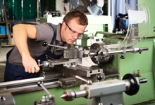 Ein Mann trägt Schutzbrille und arbeitet konzentriert an einer Drehbank in einer industriellen Werkstatt. Er trägt Arbeitskleidung und bedient die Maschine mit beiden Händen. Das Bild thematisiert den sicheren Umgang mit Maschinen im Rahmen des Arbeitsschutzes und der Prävention von Unfällen, passend zum Kontext der Unfallkasse NRW.