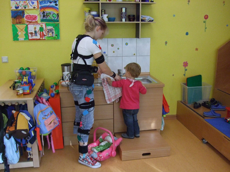 Eine Frau, die einen Bewegungsanalyseanzug trägt, steht in einer Kindertagesstätte vor einer Spielküche. Sie assistiert einem kleinen Kind, das vor dem Waschbecken auf einem Tritthocker steht. Der Raum ist bunt mit Spielzeug und Kinderkunstwerken dekoriert. Diese Szene betont die Wichtigkeit von Arbeitsschutz und Prävention bei Aktivitäten mit Kindern, wie sie von der Unfallkasse NRW gefördert wird.