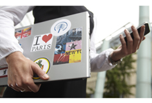 Eine Person in Business-Kleidung hält in der rechten Hand ein Smartphone und in der linken einen Laptop, der mit diversen Aufklebern versehen ist, darunter 'I ❤️ Paris' und 'New York'. Das Bild vermittelt den Kontext mobiler Arbeit und ihre Relevanz für Arbeitsschutz und Prävention, insbesondere im Hinblick auf sicheres Arbeiten unterwegs. Im Hintergrund sind moderne Gebäude und Bäume zu erkennen, die eine städtische Umgebung andeuten.