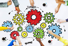 Das Bild zeigt eine Gruppe von neun Händen, die auf einen Tisch ausgerichtet sind, um mehrere bunte Zahnräder. Die Zahnräder sind rot, grün, gelb und blau und ineinander verschränkt, was symbolisch für Zusammenarbeit steht. In der Mitte des Bildes befindet sich das Wort 'TEAMWORK'. Dieses Bild passt zum Konzept von Arbeitsschutz und Prävention durch gemeinschaftliche Anstrengungen, wie sie von der Unfallkasse NRW unterstützt werden.