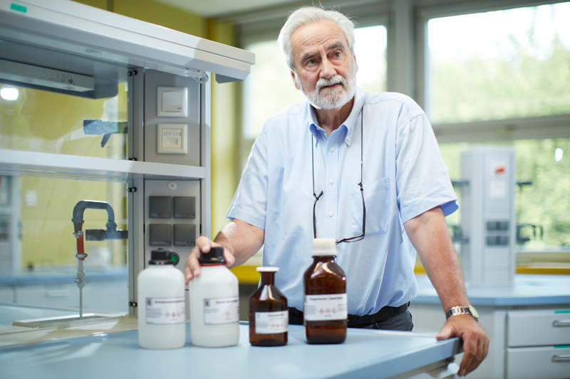 Ein älterer Mann mit weißem Haar und Bart steht in einem Labor und betrachtet mehrere Chemikalienflaschen auf einem Tisch. Er trägt ein hellblaues Hemd und eine Brille, die er um den Hals hängen hat. Die Umgebung wirkt modern und gut beleuchtet, mit Laborgeräten und Sicherheitseinrichtungen. Der Fokus liegt auf Arbeitsschutz und Prävention in der chemischen Laborarbeit, im Kontext der Unfallkasse NRW.