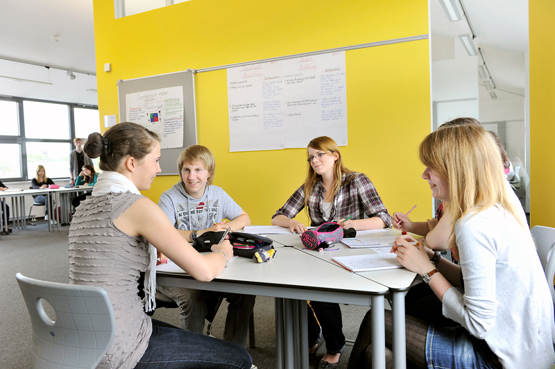 Eine Gruppe von fünf Schülern sitzt in einem modernen Klassenzimmer an zwei zusammengestellten Tischen. Sie sind in eine Gruppenarbeit vertieft, wobei einige Notizbücher und Stifte vor sich liegen. Der Raum ist hell und die Wände sind gelb gestrichen. An der Wand hängt ein Whiteboard mit Notizen. Die Atmosphäre wirkt konzentriert und kooperativ, was auf Teamwork und Lernfokus hindeutet. Kontextualisiert für Arbeitsschutz und Prävention im Bildungsumfeld passend zur Unfallkasse NRW.