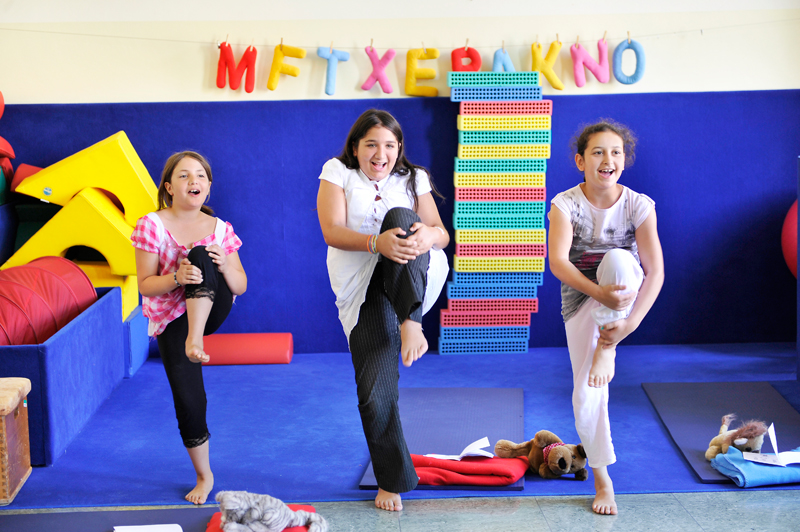 Drei lachende Mädchen in legerer Kleidung heben synchron ein Bein in einem Raum mit bunten Matten und Spielzeug. Im Hintergrund hängen bunte Buchstaben an der Wand, die unleserlich angeordnet sind. Die Szene deutet auf spielerisches Training zur Förderung von Gleichgewicht und Koordination hin, das im Kontext von Präventionsmaßnahmen und Arbeitsschutz für Kinder steht.